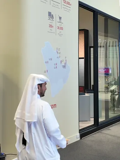 A visitor in a white thobe and ghutra stands beside the wall of the ABYAT stand.