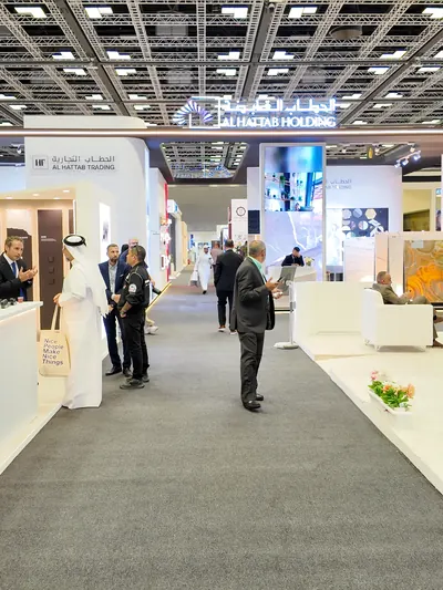 Visitors walk down a carpeted aisle toward the Al Hattab Holding stand, under the lit lattice ceiling of the hall.