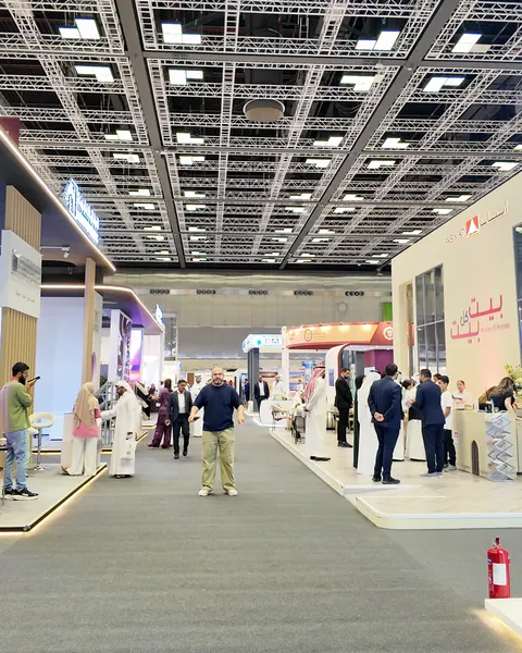 A wide exhibition hall under a white lattice ceiling, with visitors gathered along a carpeted aisle between the stands.
