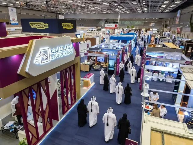 Visitors in traditional Qatari dress walking along a carpeted aisle between exhibition stands in a large hall, with the Al Hattab Holding stand in the foreground.