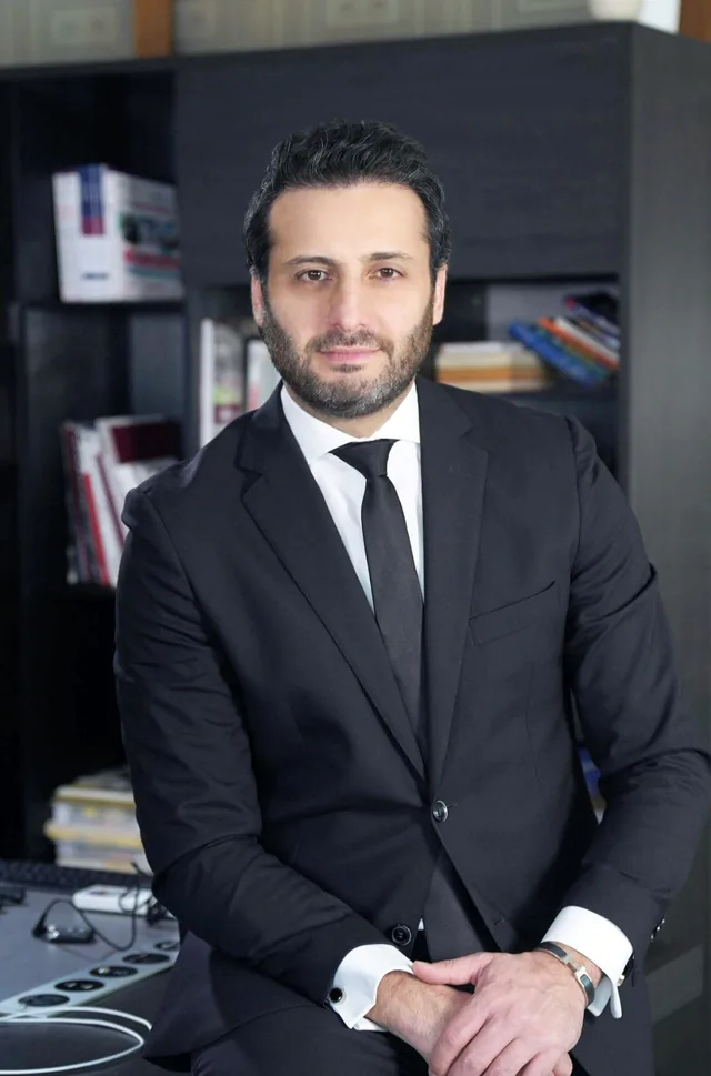 Rawad Sleem, Co-Founder and General Manager of NeXTfairs, in a dark suit and tie in front of a bookshelf.